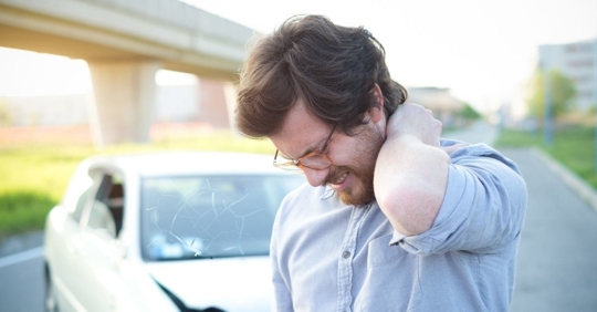 man holding his neck in pain standing in front of a crashed car