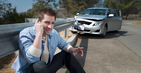man on the phone sitting on the curb in front of a crashed car